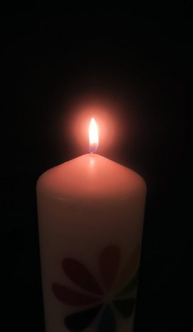 Dunkelheit, darin eine weiße, erleuchtete Kerze mit dem Unitariersymbol (eine stilisierte Blüte mit regenbogenfarbigen Blättern)