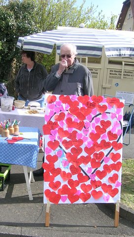 Foto eines weissen Posters auf die viele kleine rote und rosafarbene, beschriftete Herzen geklebt sind.  Im Hintergrund stehen zwei Personen.